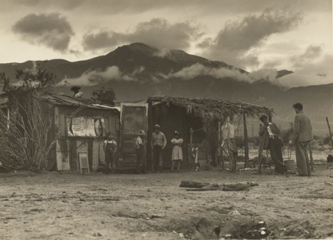 A ramshackle patchwork structure sits in front of some nearby hills. 4 children, likely farm workers, are standing around. An adult male immigrant farm worker is talking to a white man on the right of the picture.  The white man is presumbly Paul Taylor.