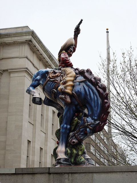 A sculpture of a blue horse bucking while a brown-skinned vaquero, or cowboy, proudly rides the horse. The statue dominates the center of the photograph, in front of an imposing white marble building with stylized columns.