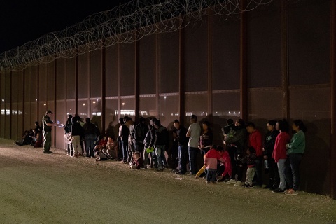 A US Customs and Border Protection agent is examining papers in front of a line of men, women and children. They are all in front of a tall wall with concertina wire.  It is night.