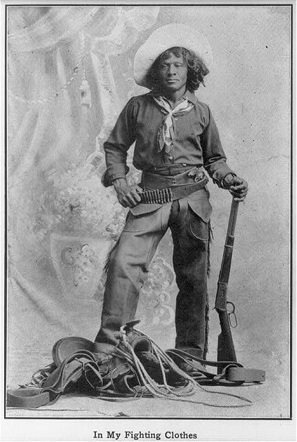  Nat “Deadwood Dick” Love poses in front of a studio backdrop in this black and white photograph. He wears a cowboy hat over his shoulder-length hair, a button-down shirt, and chaps. He puts one hand on his hip and rests the other on his rifle. 