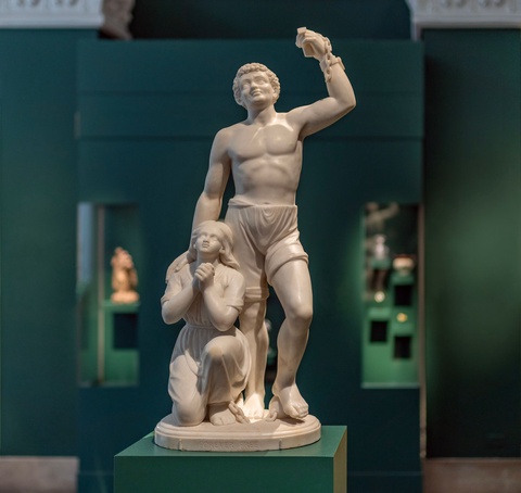 A photograph of a white marble sculpture in a green museum gallery. The sculpture consists of a standing shirtless man wearing trousers. He gazes upward while raising one fist in the air, dangling a broken shackle. He rests his hand on the shoulder of a  woman with long hair in a simple dress, who kneels next to him. She gazes upward, her hands clasped as if in prayer. 