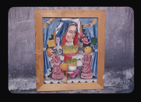 A photograph of a painting in a light-colored wood frame leaning against a gray concrete or stone wall. The painting shows an altar with a portrait of the crying, blue-veiled Virgin Mary, sprouted wheat, an unlit candle, roses, and small figurines of angels and a horse.