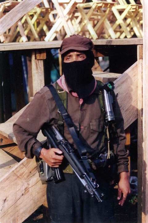 The photograph features a brown-skinned person wearing dark-colored clothing, with a balaclava covering all of their face except their eyes. They have a gun strapped across their chest and stand in front of simple wooden stairs.