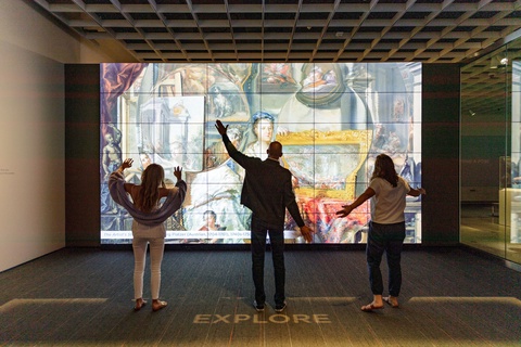 Three museum visitors are facing a large-scale illumination with their hands in various gestures becaues they are interacting with a gesture-based ArtLens Display at the Cleveland Museum of Art.   