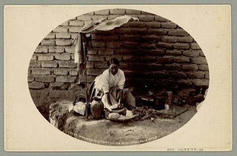 An oval grayscale photograph on a background of aged buff paper, which has the appearance of a photo in an album. A woman with a medium-dark skin tone and dark hair gazes at the camera with a serious expression as she kneels on the ground grinding corn for tortillas. She is surrounded by cooking implements and works on bare earth beside a stone wall.