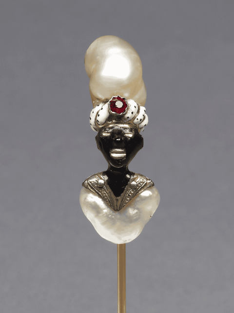 The head of the gold pin is in the form of an African with an enameled face. Both his turban and chest are baroque pearls. The rim of the turban is in white enamel with diagonal rows of black dots and is set with a red paste.