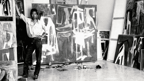 Artist Wilfredo Lam standing in his studio, gazing at the camera, surrounded by paintings leaning on the walls.
