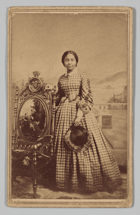 A sepia tone carte-de-visite of a young woman, captured at the Ball & Thomas Photographic Art Gallery in Cincinnati, OH. Standing in front of an elaborate outdoor backdrop, a dark-haired woman poses before the camera, slightly smiling, dressed in a floor length checkered dress with her hair styled in a chignon. She holds a feathered dark hat in her proper left hand, with her proper right hand resting on the high back of an ornate wooden chair. The front of the chair, which faces the camera, features a patterned fabric seat, scroll and floral carvings, and an oval painting of a man and child on the chair’s back.