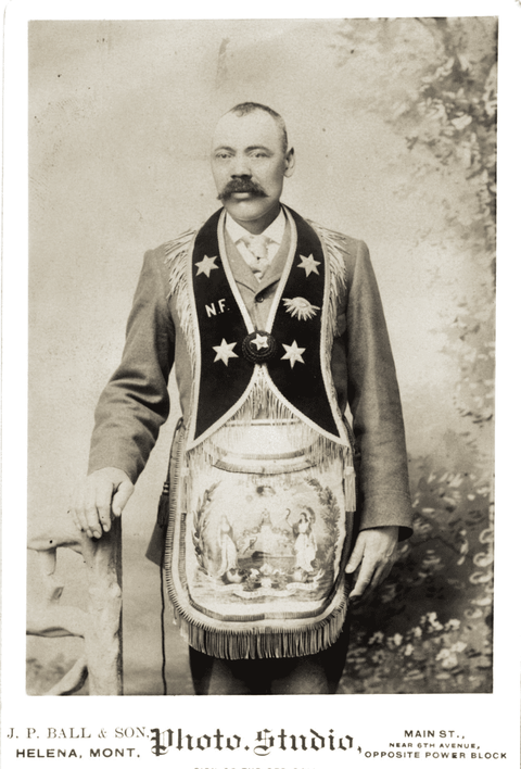 Black and white portrait of a mustachioed man wearing fraternal organization regalia in front of a nature scene backdrop. 