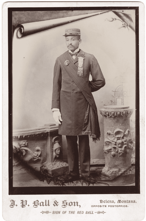 Studio portrait (full body) of William C. Irvin, a black man with a mustache, wearing a uniform. Irvin became a policeman in Helena, Montana, in 1888. He wears what may be his uniform, including a hat, and a ribbon for the Grand United Order of Odd Fellows in America, the largest African American fraternal group in the country.