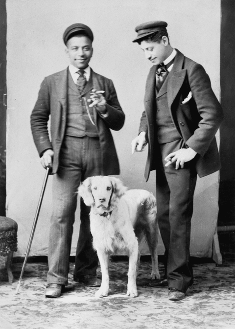 Full body portrait of a two unidentified African-American men wearing hats with a dog. Man on left holding a cigar in left hand and cane in right is standing near a stool and has a chin attached to his vest. Man on right wearing a hat and is looking down pointing his right finger and holding a cigar in his left hand.