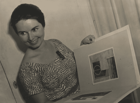 A black and white photograph of a smiling woman with short hair holding open a large bound portfolio