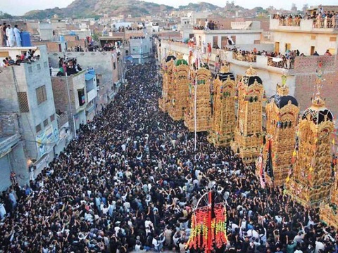 A street densely packed with people, most wearing black or dark colors. A row of tall, ornate gold paper and bamboo temples are being carried by the crowd.