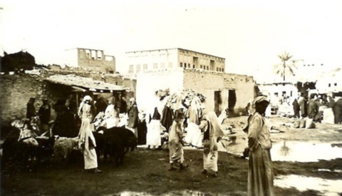 A black and white photograph of a wide, open, market, some people standing in the foreground. 
