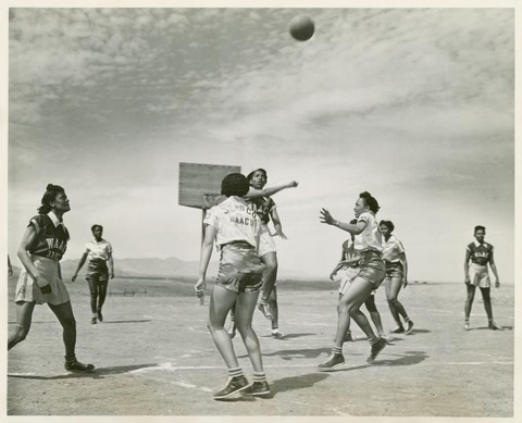 A black-and-white photograph depicts eight African American women wearing short sleeve shirts, skirts and leather athletic shoes playing basketball. There appear to be two teams, distinguished by white and black shirts.