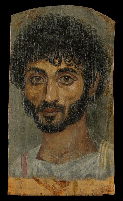 A portrait of a man with dark and thick curly hair. He has a beard and is wearing a white garment.