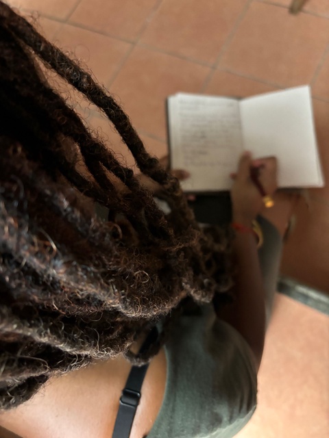 A color photo depicts the author, Mi’Jan Celie Tho-Biaz, writing in a leather bound journal, holding a pen. The camera has focused the photo on the different strands of her loc hair, black bra strap and green dress. Her face is obscured. 