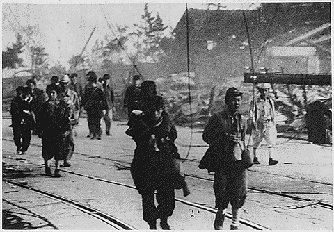 A group of people move toward the photographer with panicked expressions against a backdrop of rubble. The photograph is black and white. 