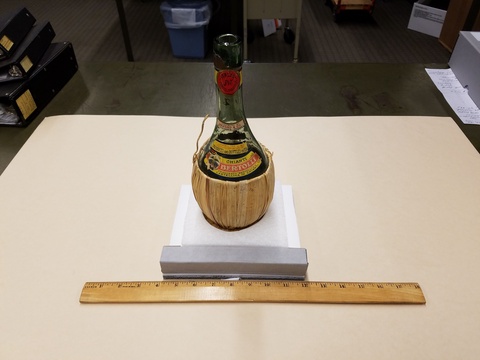 An empty wine bottle sits on a square pedestal on a white table. A ruler placed in front of the pedestal shows that the bottle is about four inches wide. The wine bottle is made of greenish glass with a red seal and a basketweave fiver base.