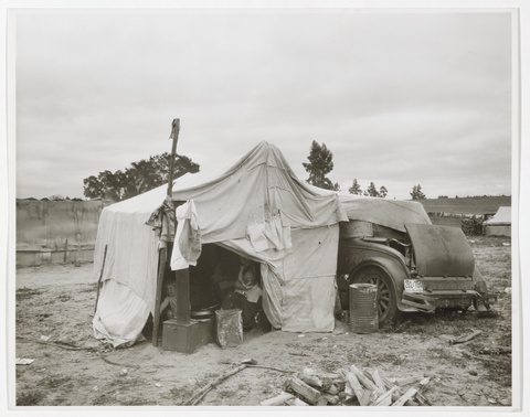 A black and white photograph depicts a temporary shelter made of sewn-together tarps propped up by poles and draped over a dilapidated car. 