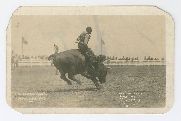 A worn, black-and-white picture postcard of a Black cowboy riding a bucking bull in an open field. Handwritten text on the bottom of the image reads: “California Rodeo Salinas 1919. Official photo #45 by Marcell.” 