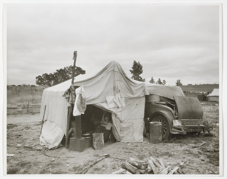 A black and white photograph depicts a temporary shelter made of sewn-together tarps draped over a dilapidated car. On the left side of the entrance, a child peers out; also in the shadows on the right, a woman sits holding an infant on her lap.