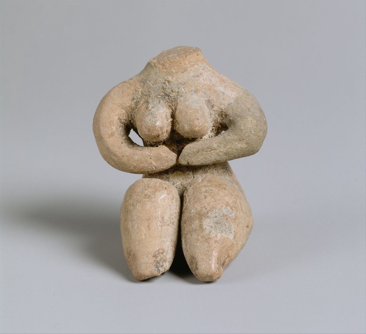 A small, ceramic, Halaf figure with traces of paint. The figure is headless but has prominent breasts framed by the arms which are folded underneath. 