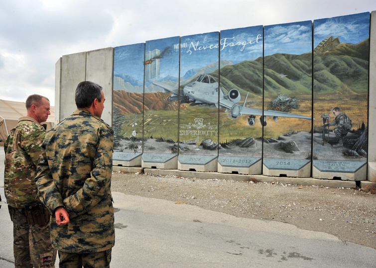 Two men in army camouflage stand outside under gray skies, looking at a mural painted on a series of segmented concrete blast walls. 