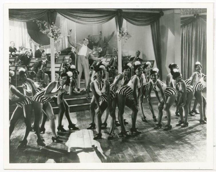 A black and white photograph of Cab Calloway with his dancers at the Cotton Club. Calloway stands in the middle, onstage with his arms outstretched. He holds a conductor's baton in his right hand. His band is seated, playing instruments behind him. 