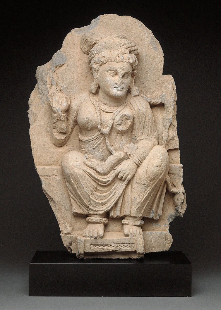 A Gandharan sculpture of the goddess, Hariti, seated and holding a child in her left arm. Her right hand, now partially destroyed is raised in a mudra. She is robed and wears barcelets, anklets necklaces and large earrings. 