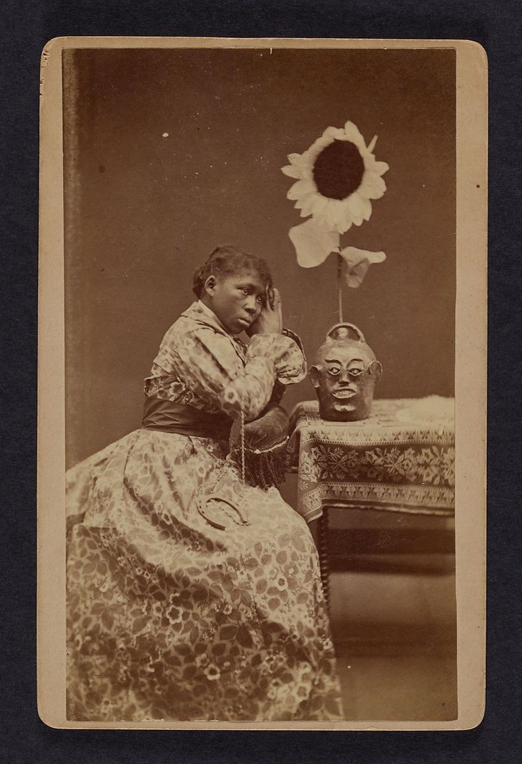 A photographic portrait by James A. Palmer of a young Black woman wearing a floral dress. She is seated next to a table. Her arms rest on the chair's back and her hands are clasped in a prayer pose.