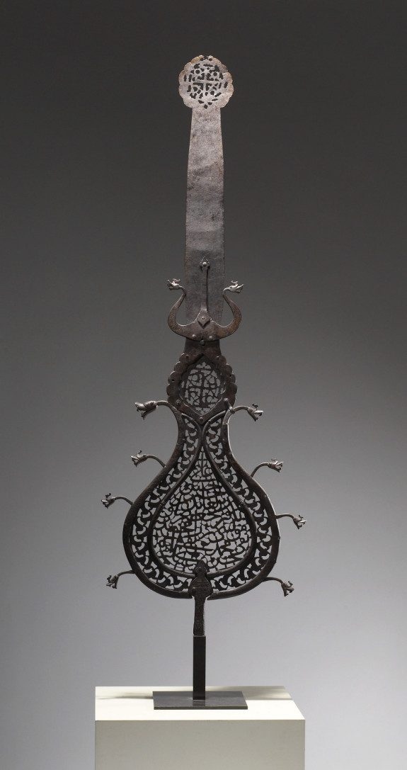 An Iranian, metal 'alam from the 17th century. The center and top contain calligraphy. Five dragon heads protrude from each side of the body.