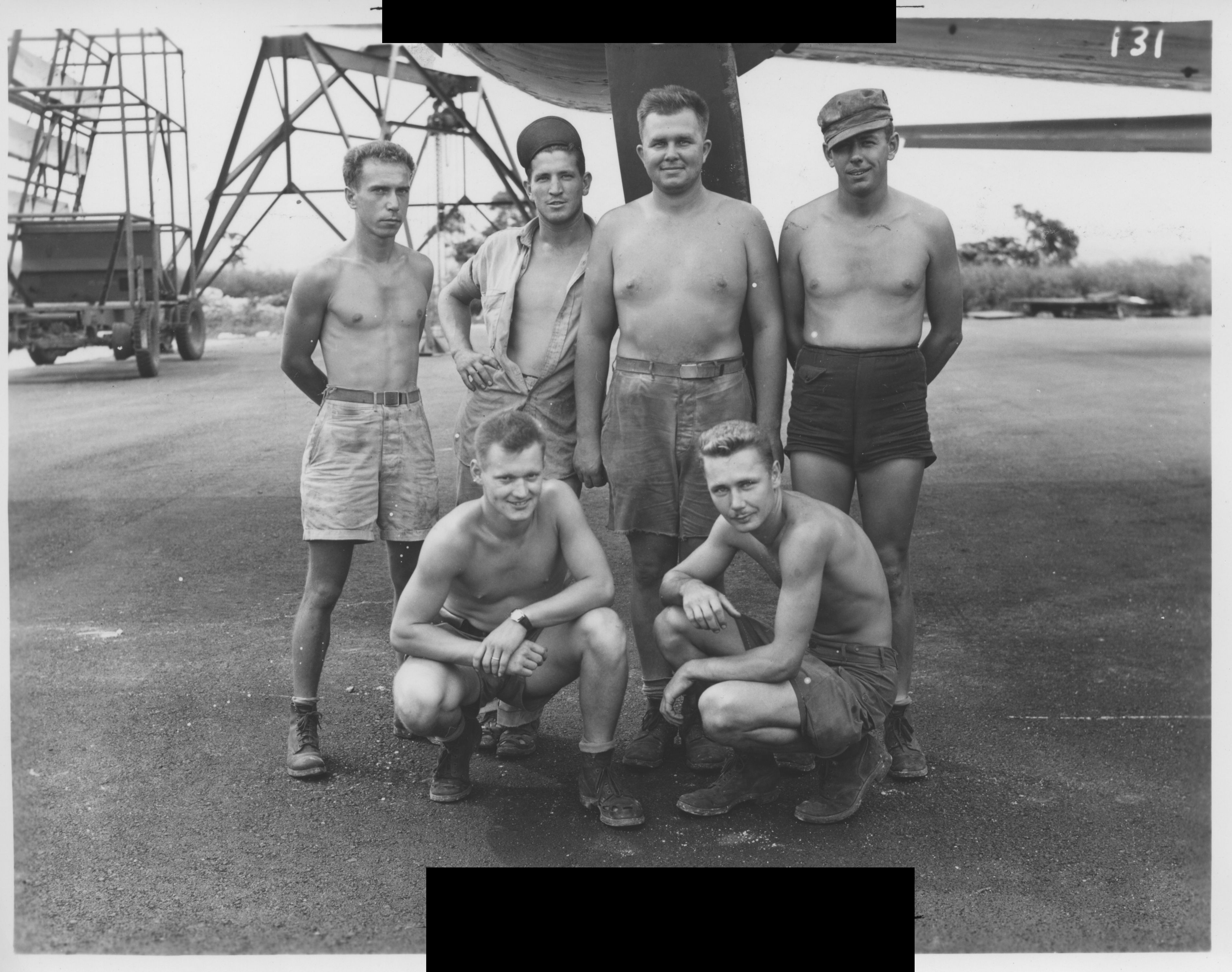 A group of five shirtless, light-skinned men pose outside against a background of heavy machinery. The men have close-cropped hair and most of them smile proudly. The photograph is black and white.