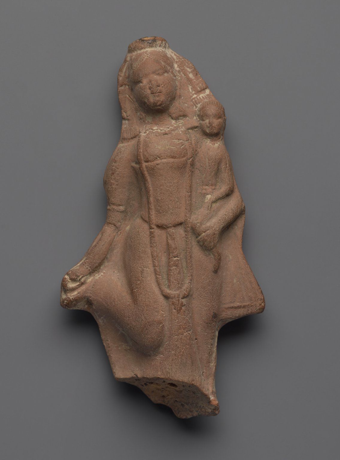 A small terracotta plaque of the goddess, Hariti, holding a child in her left arm. She wears a long necklace and armbands and holds her right food in her right hand.