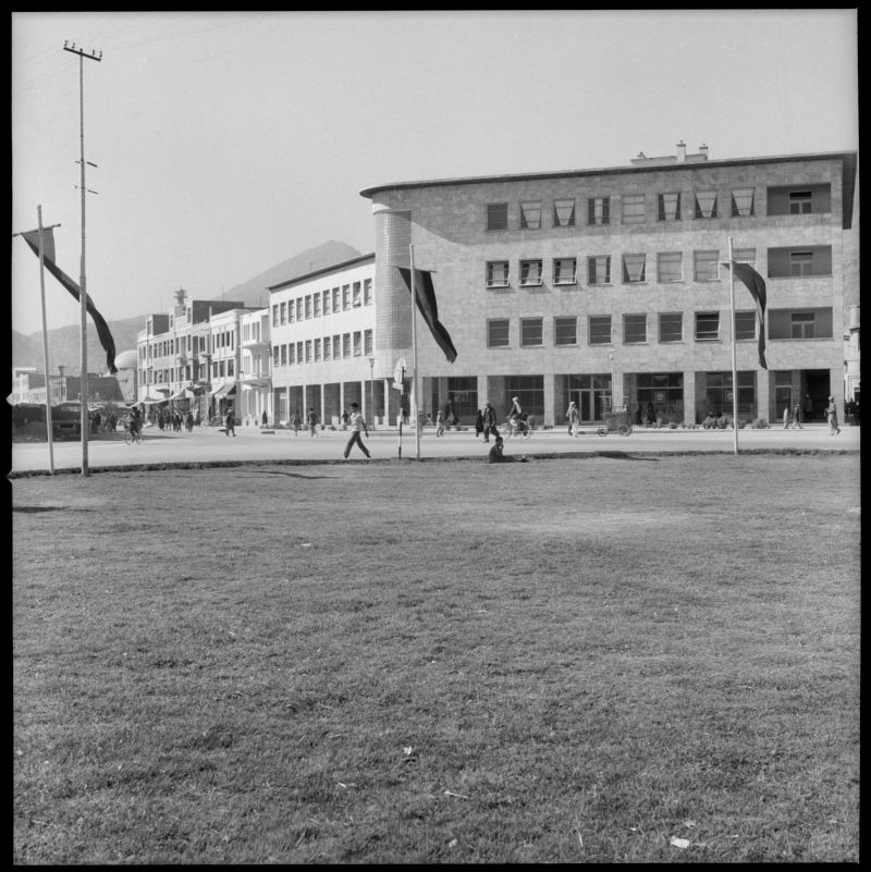 A black and white photograph of commercial buildings taken from a lawn. The grass fills the lower half of the picture. People pass by on the sidewalks and flags wave in the wind.