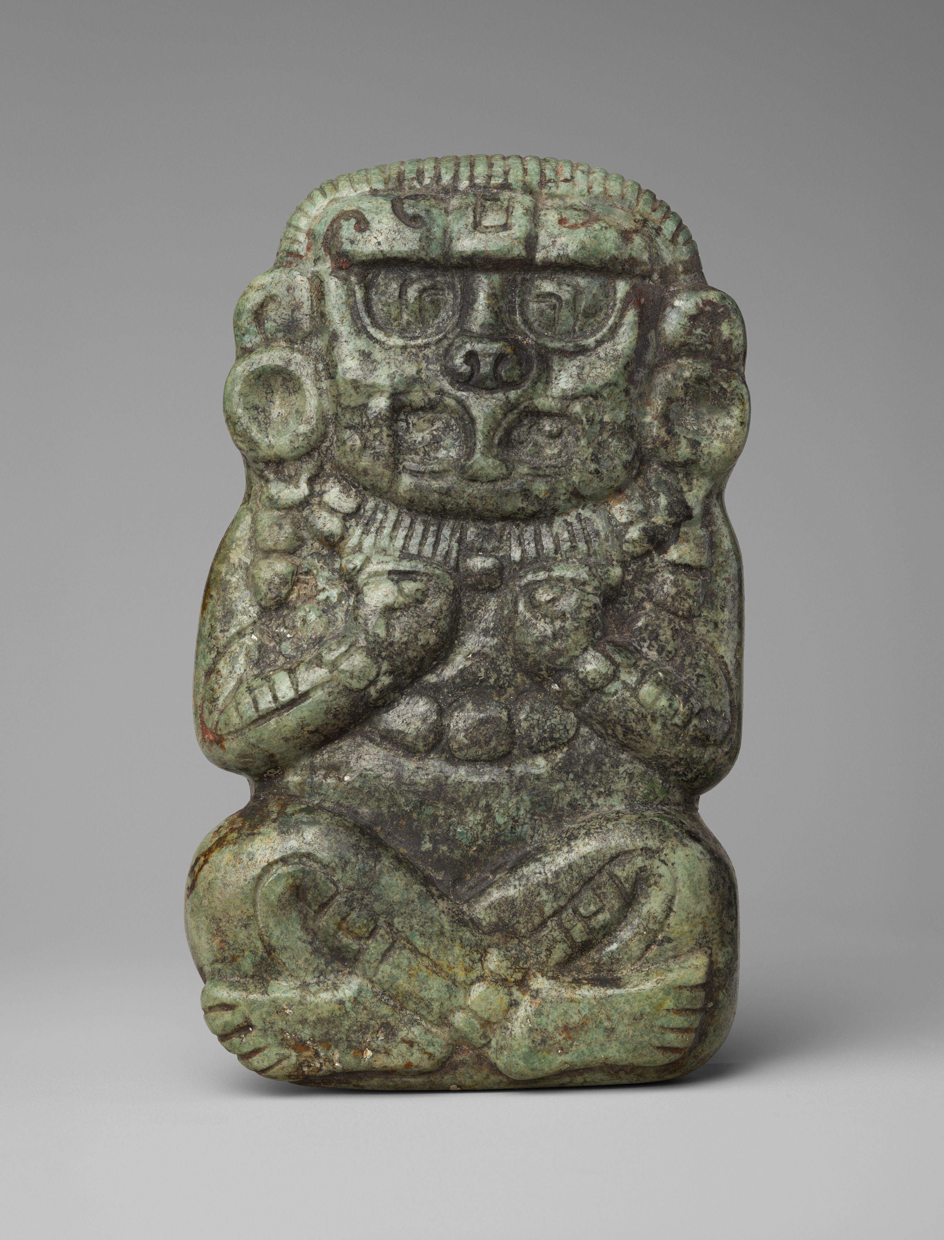 A Mayan jade carving of a deity. The figure is seated crosslegged with its hands up to its chest. Their face has avian features. The figure wears ear gauges, necklaces, bracelets, and anklets.