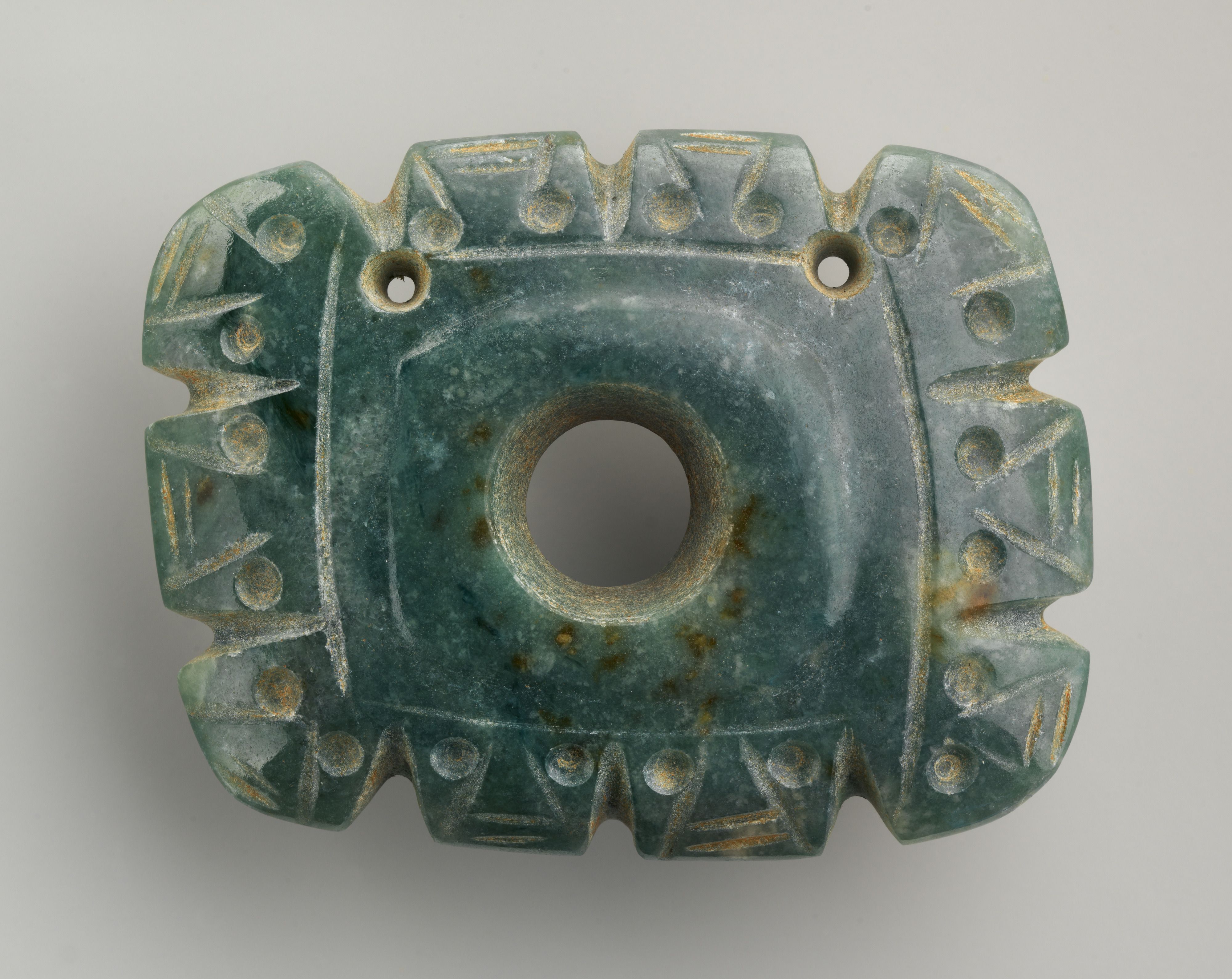 A small, rectangular, blue green jade pendant. The pendant has a large hole in the middle and two smaller ones above it. Carved heads with large eyes border the pendant.
