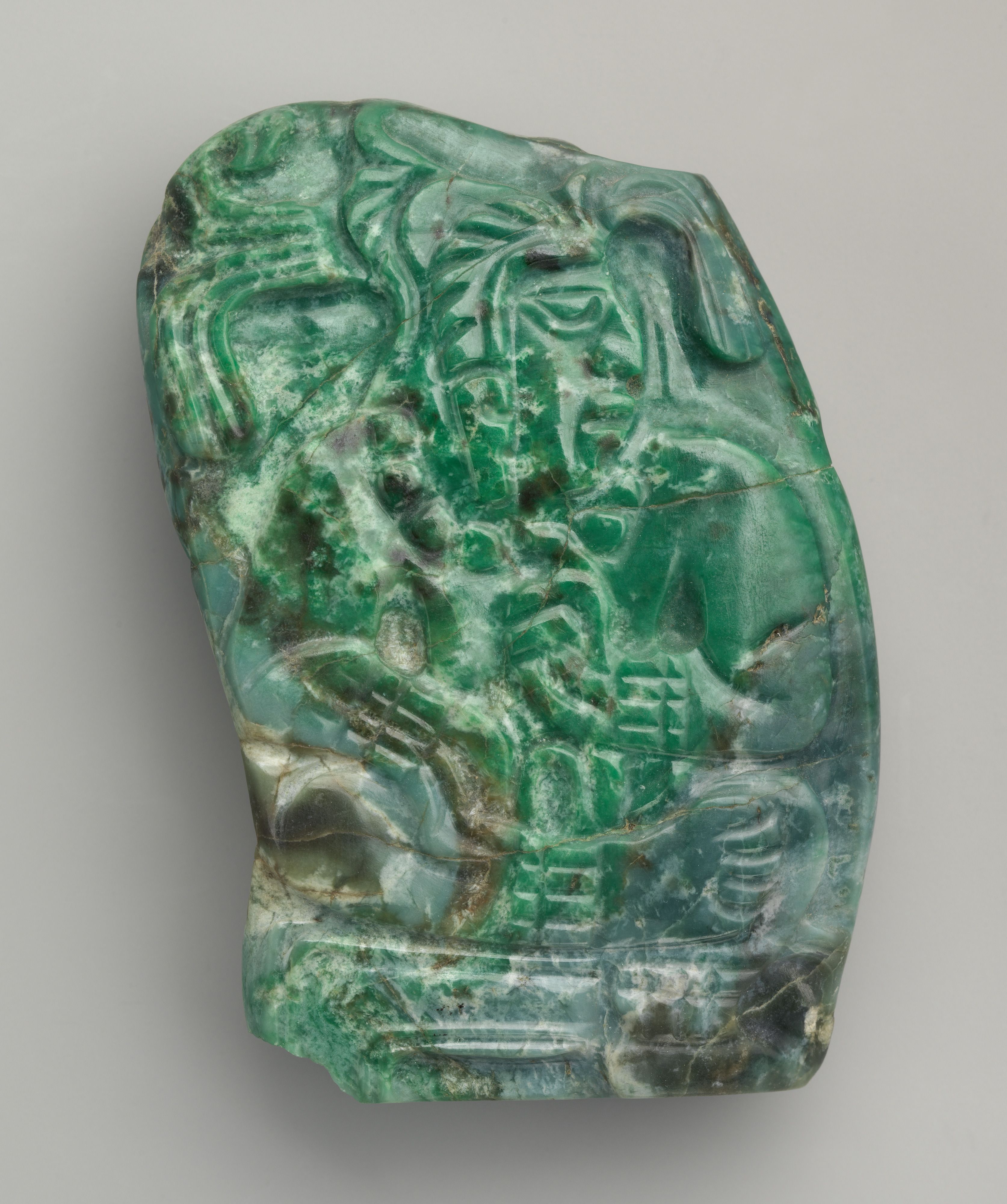 A small jade pendant featuring a lord seated crosslegged on a dais. The figure turns his head to his left and wears a feathered headdress. He wears a necklace, bracelets, and anklets. His left hand is raised to his chest.