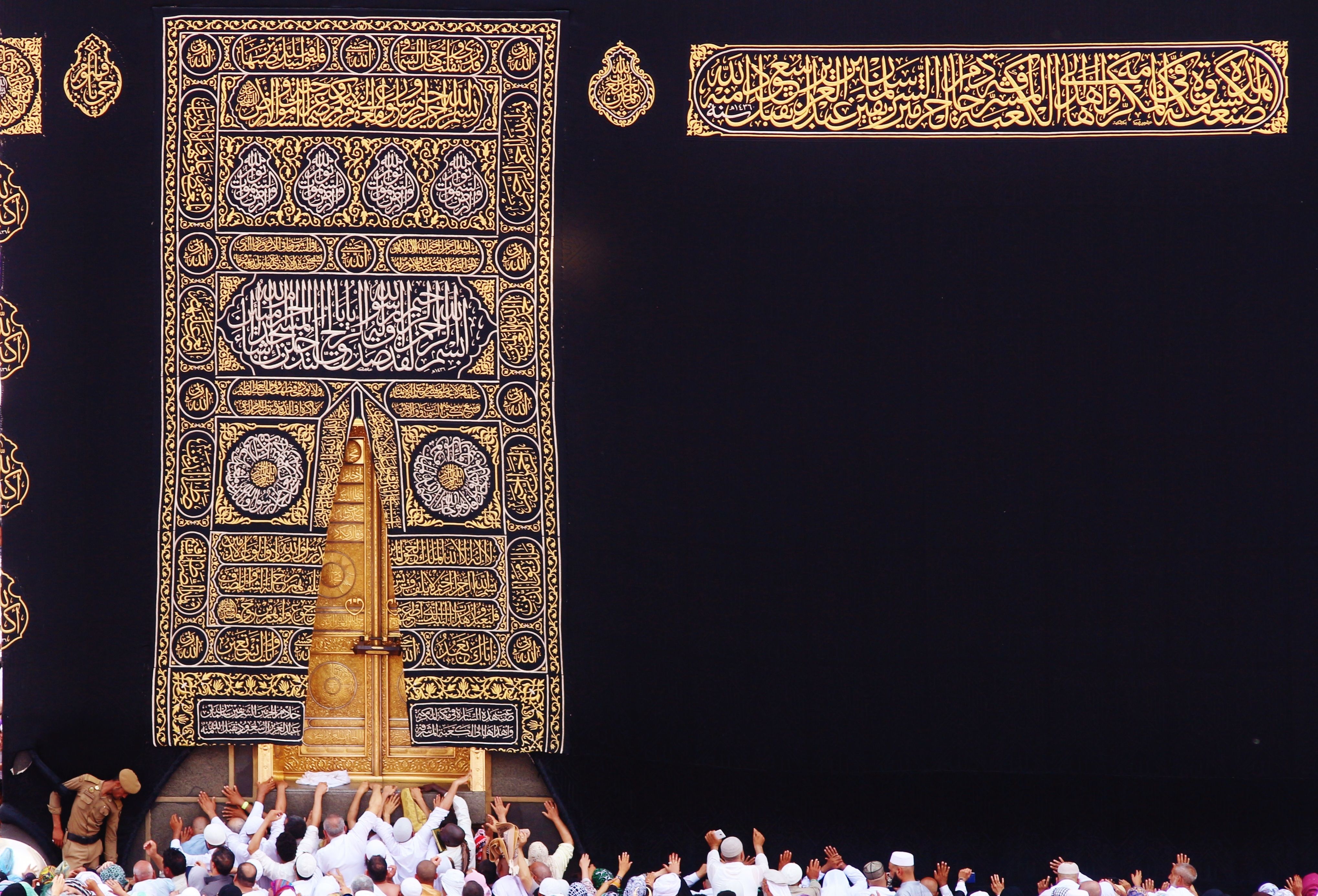 A large black textile densely embroidered in places with gold and silver Arabic calligraphy forming geometric designs, including the shape of a door, partially covers an actual door. Worshippers dressed in white gather along the bottom edge of the image.