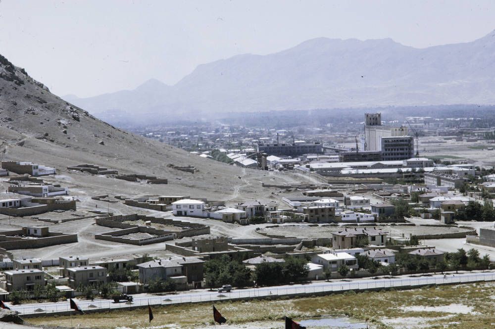 A bird’s eye view of Kabul’s cityscape, including the tower of a bakery and a steep slope. A mountain range is visible in the background.