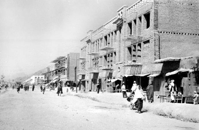 A black and white photograph taken facing one side of a street in Kabul, Afghanistan. The buildings are newly constructed and people pass by on the side of the roadway.
