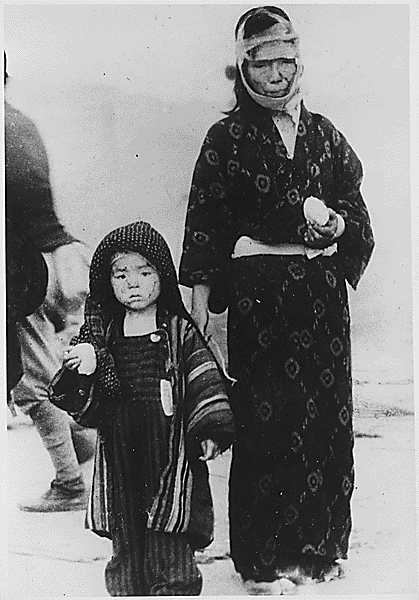 A woman stands next to a small child, facing the camera. The woman wears a black kimono with a simple pattern and has bandages on her head. Both the woman and child carry rice balls. Their expressions are grim. 